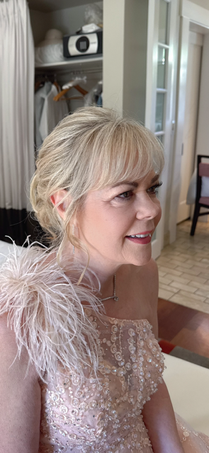 Blonde woman in a pink feathered dress smiling indoors near a mirrored cabinet.
