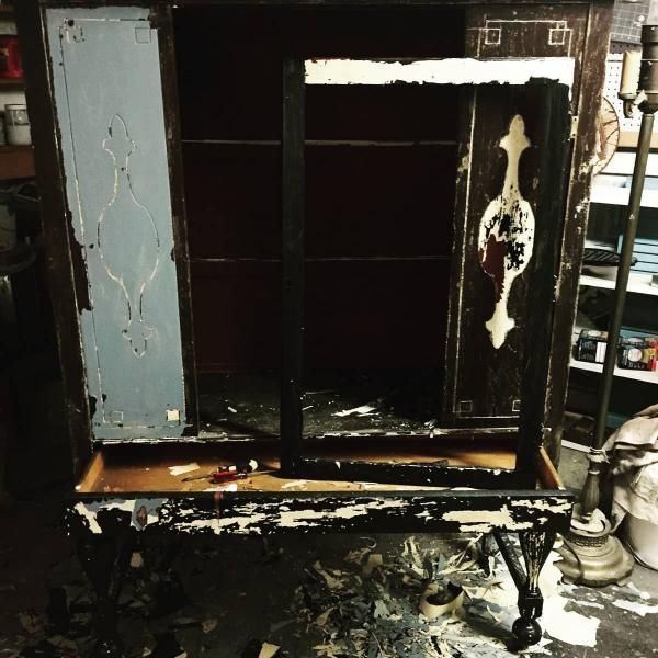 Old black cabinet with a chipped white door and worn paint, sitting on a cluttered floor.