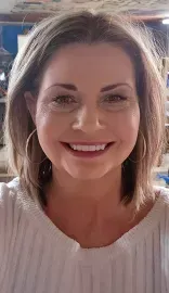 Smiling woman in a white top, seated indoors with warm light in the background.