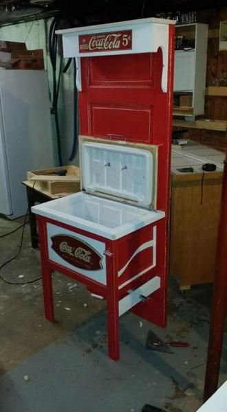 Red Coca-Cola upright cooler with open top and bottom compartments in a workshop or storage area.