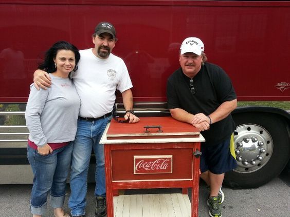 Three people posing beside a red Coca-Cola cooler in front of a red truck.