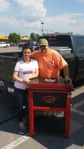 Two people stand beside a red-and-black grill in a parking lot under a cloudy sky.
