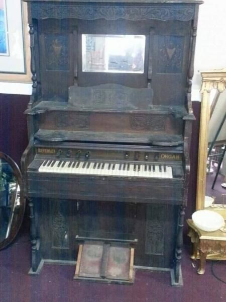 Old upright piano with ornate wood cabinet in a room, with a small bench and framed artwork nearby