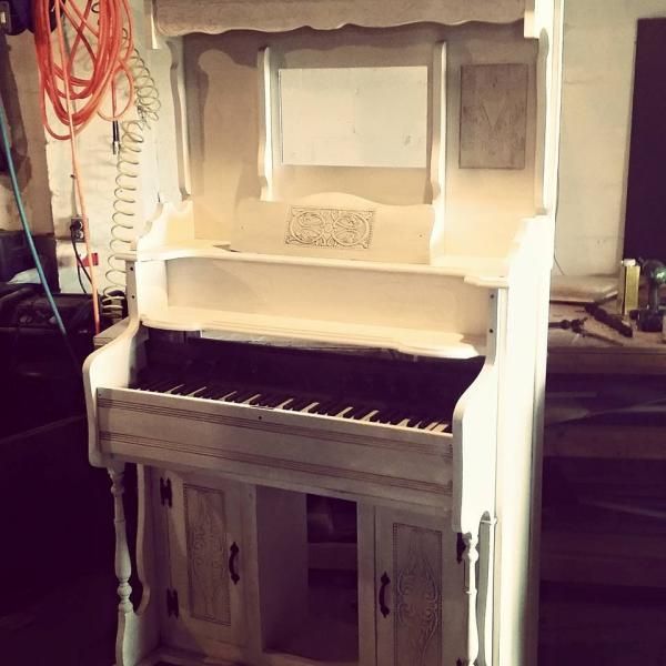 White upright piano or keyboard-style cabinet in a workshop, with a dark keyboard and storage doors below