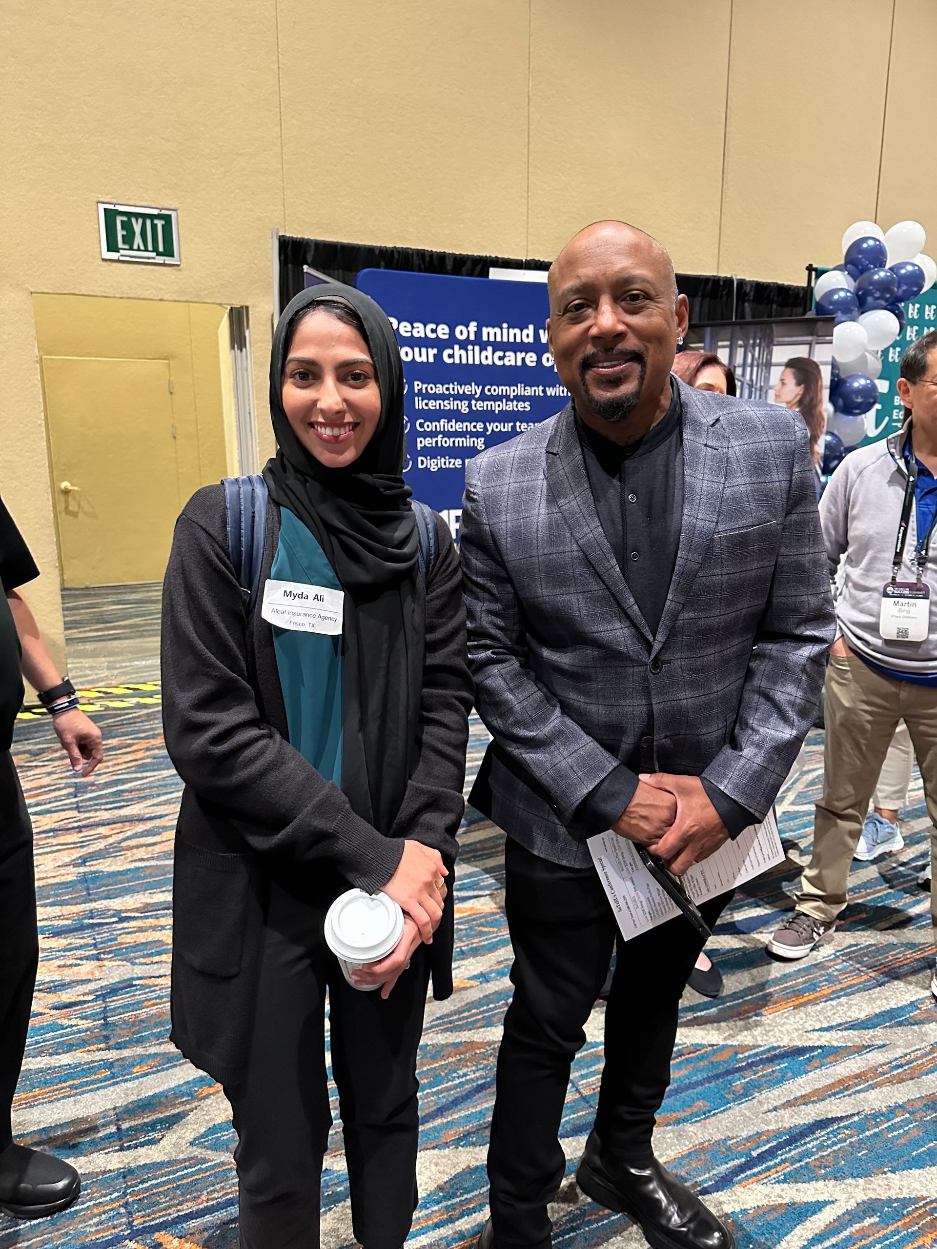 Woman in hijab and man in suit pose together indoors, smiling, near a conference display.