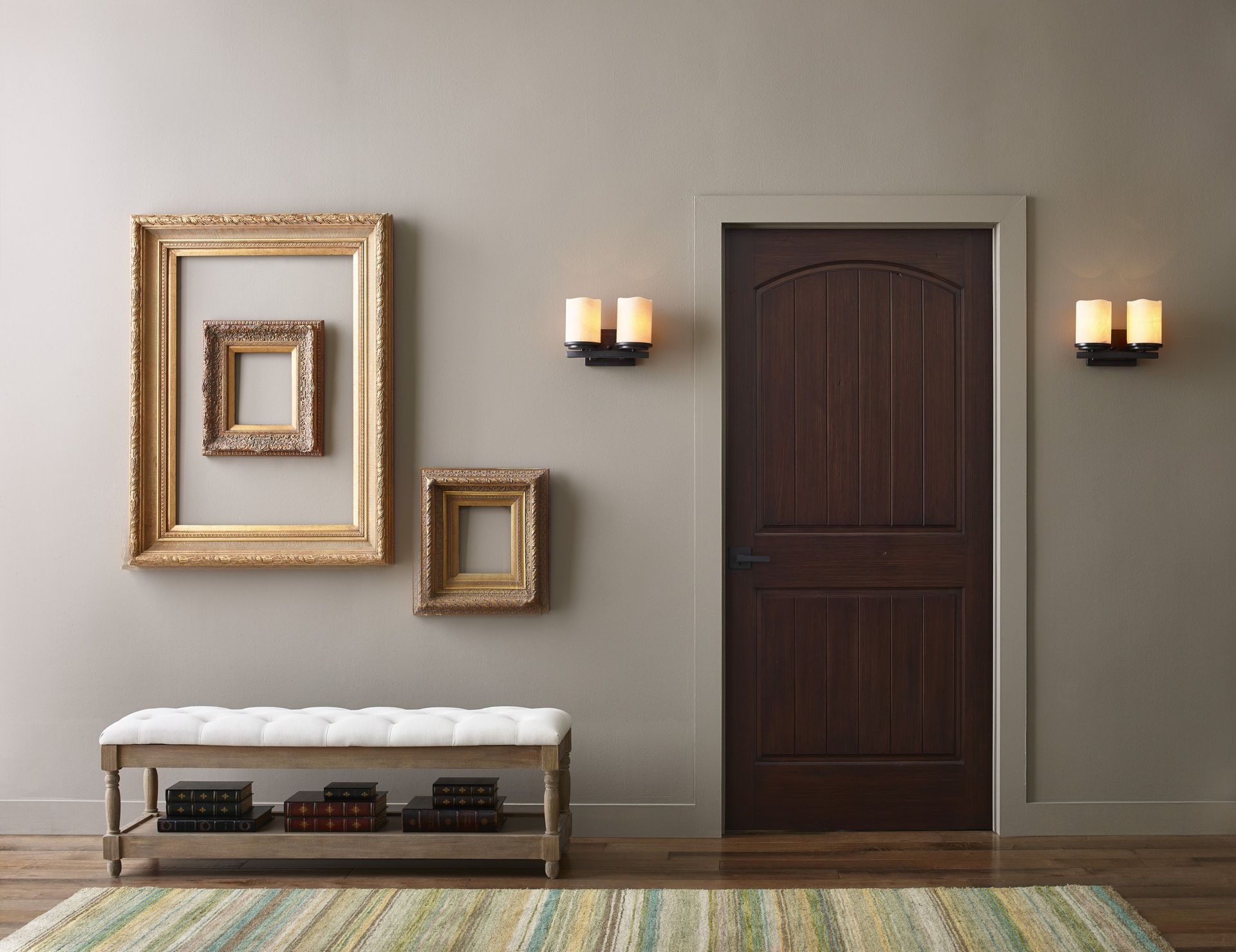 Hallway with a wooden door, three empty ornate gold frames, a bench, and sconce lights.
