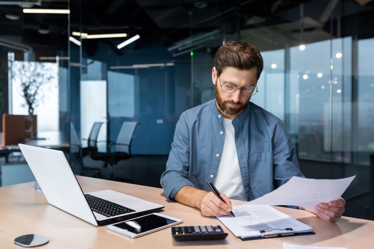 analisando-dados Um homem está sentado em uma mesa com um laptop e papéis.