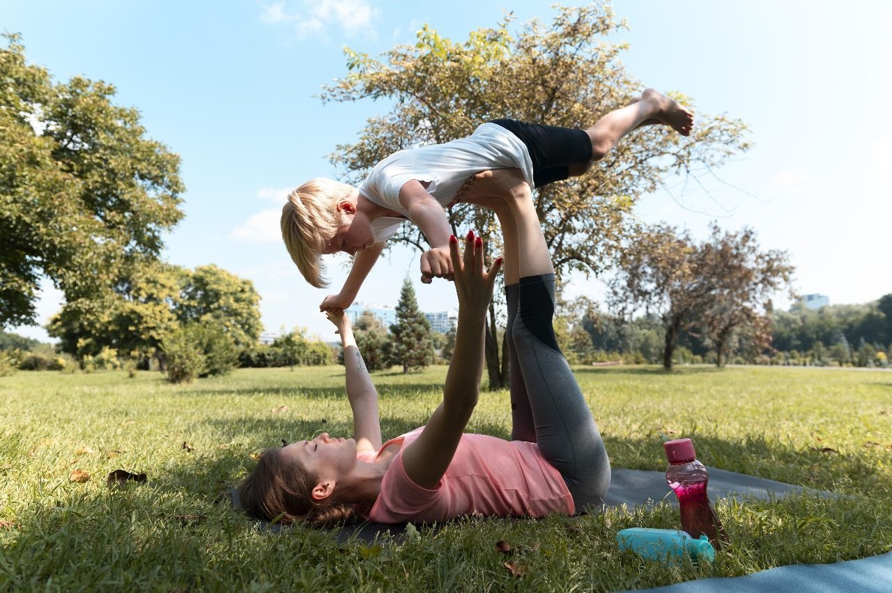 familia-em-area-verde Uma mulher segura uma criança no ar enquanto faz ioga em um parque.