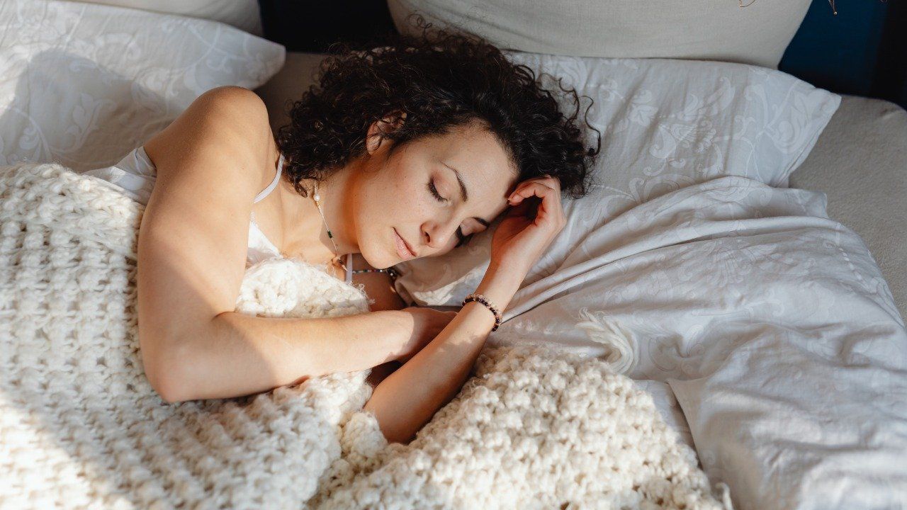 mulher-dormindo Uma mulher está dormindo em uma cama com lençóis e travesseiros brancos.