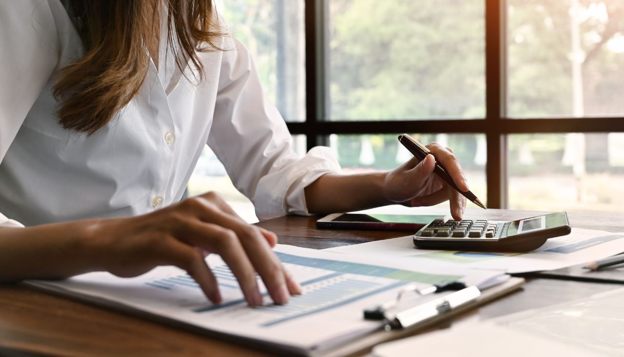 pessoa-analisando-dados Uma mulher está sentada em uma mesa usando uma calculadora e uma caneta.