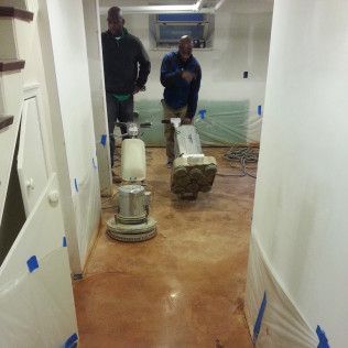 Two workers using floor grinders in a hallway, with plastic-covered walls and taped-off doorways.