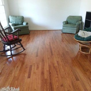 Empty living room with hardwood floors and two green armchairs by the walls, plus a rocking chair and wicker chair.