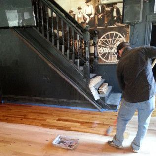 Man standing in a room beside a black staircase, looking at the floor near a small paper on wooden boards.