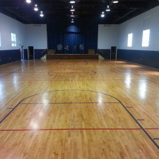 Empty indoor basketball court with wooden floor, blue walls, and a stage at the far end