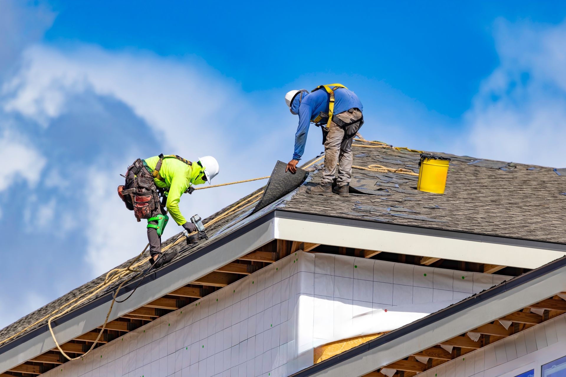 Workers repairing a roof on a house, with one in a bright green jacket and another in blue and yellow gear.
