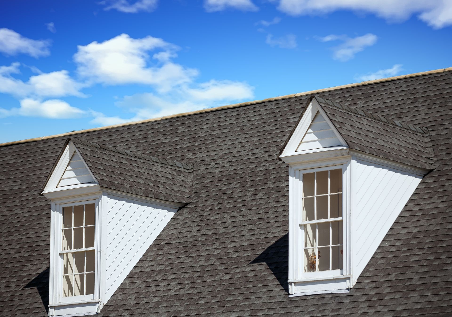 Two white dormer windows on a dark shingled roof under a blue sky