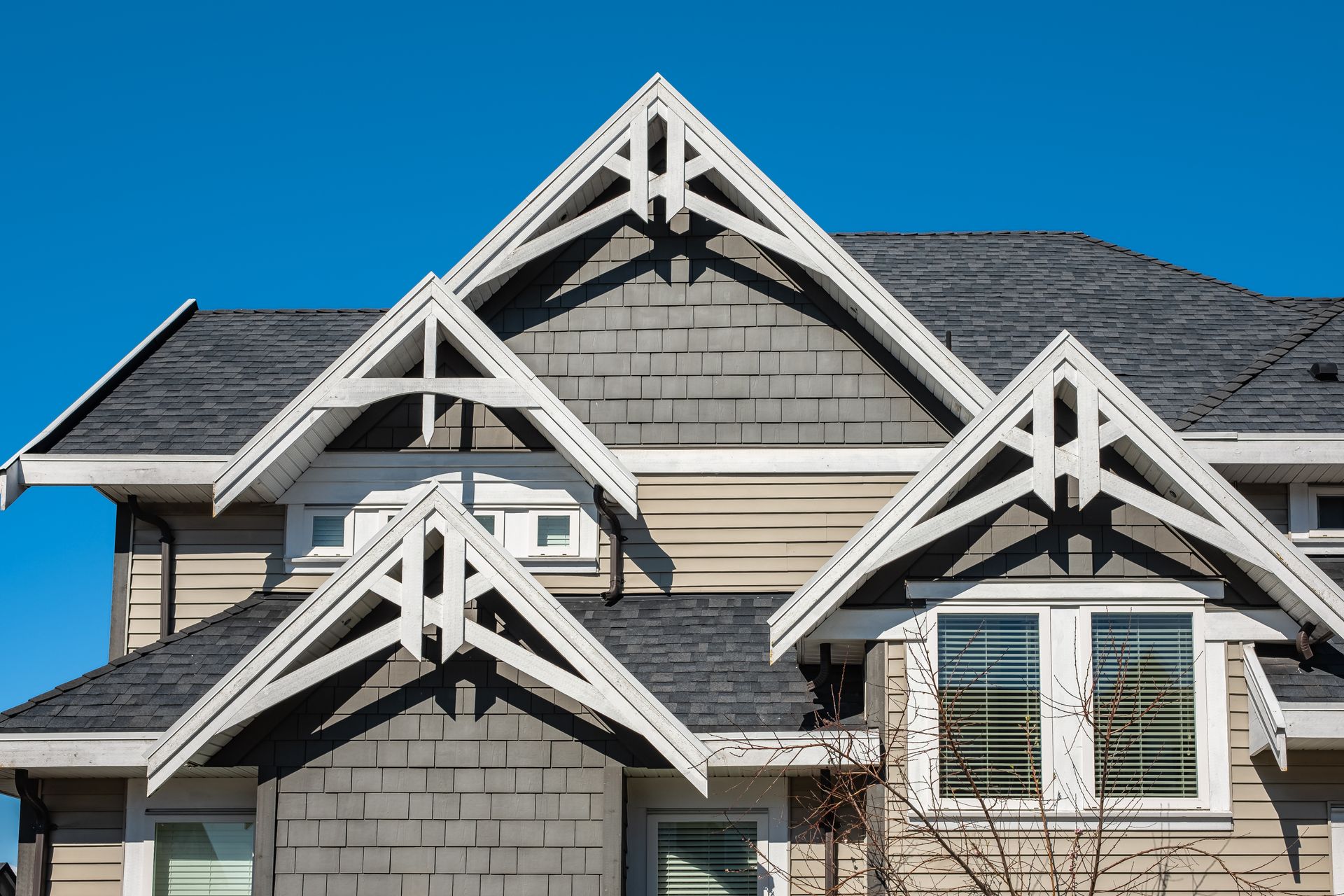 Front of a beige house with dark shingled roof and decorative white gables against a clear blue sky