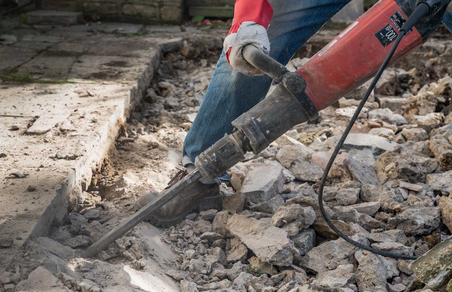 Person in blue jeans using a red jackhammer to break up concrete outdoors.