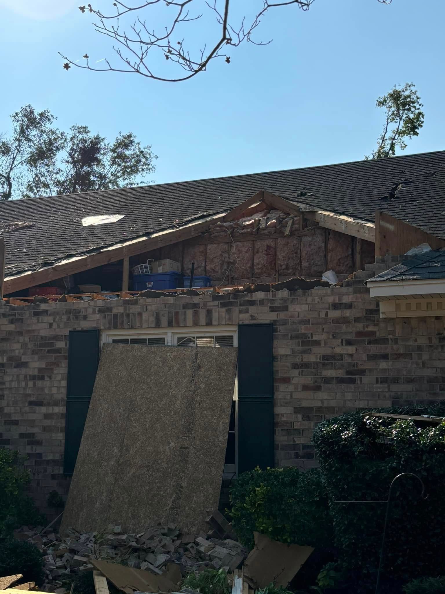 House with roof damage, exposed framing, brick exterior, blue sky, debris.