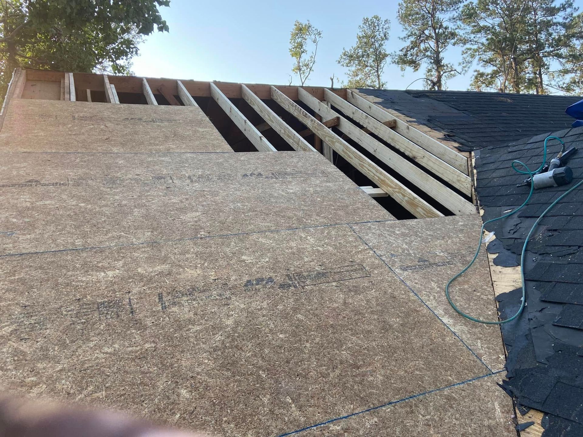 Construction of a roof. Wood beams and sheathing are exposed. A nail gun and shingles are visible on the finished section.