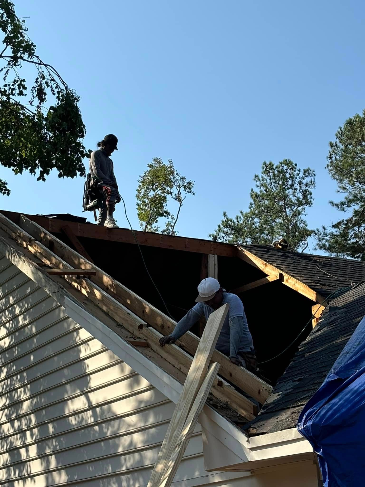 Two roofers repairing a roof, one on the roof's peak, the other near the edge; blue sky backdrop.