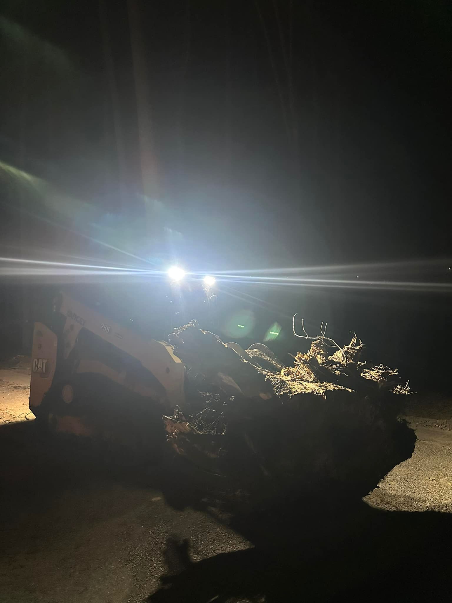 Skid steer loader illuminated by bright headlights at night, pushing material on a dark surface.