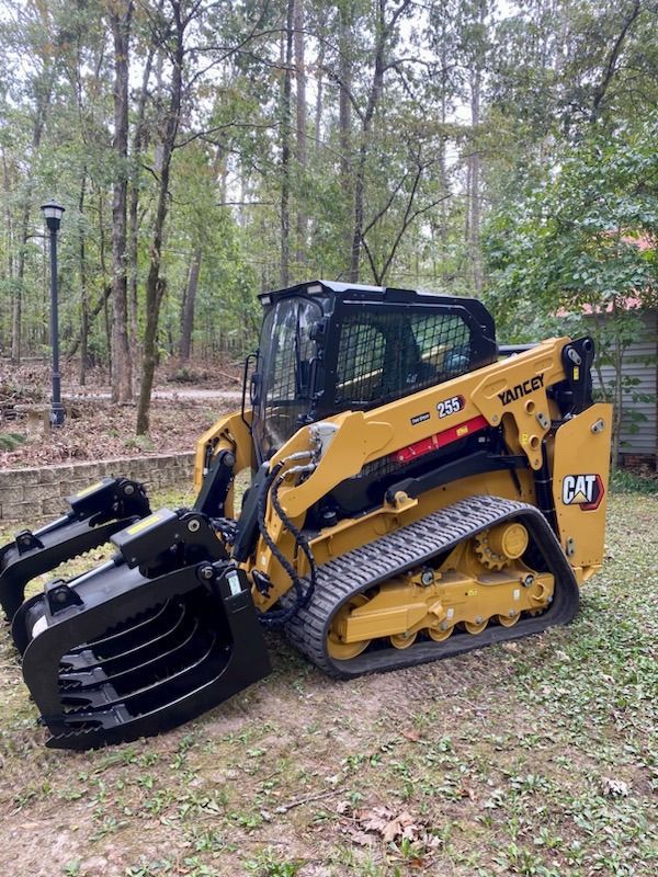 Yellow Caterpillar compact track loader with grapple attachment in a wooded area.