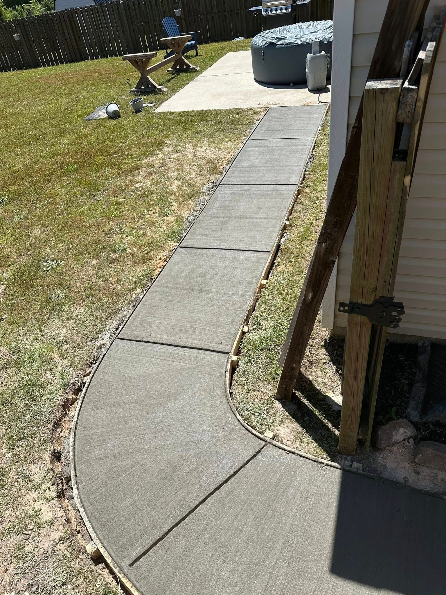 Concrete walkway curving to patio. Grass on either side, hot tub and wooden structure nearby.