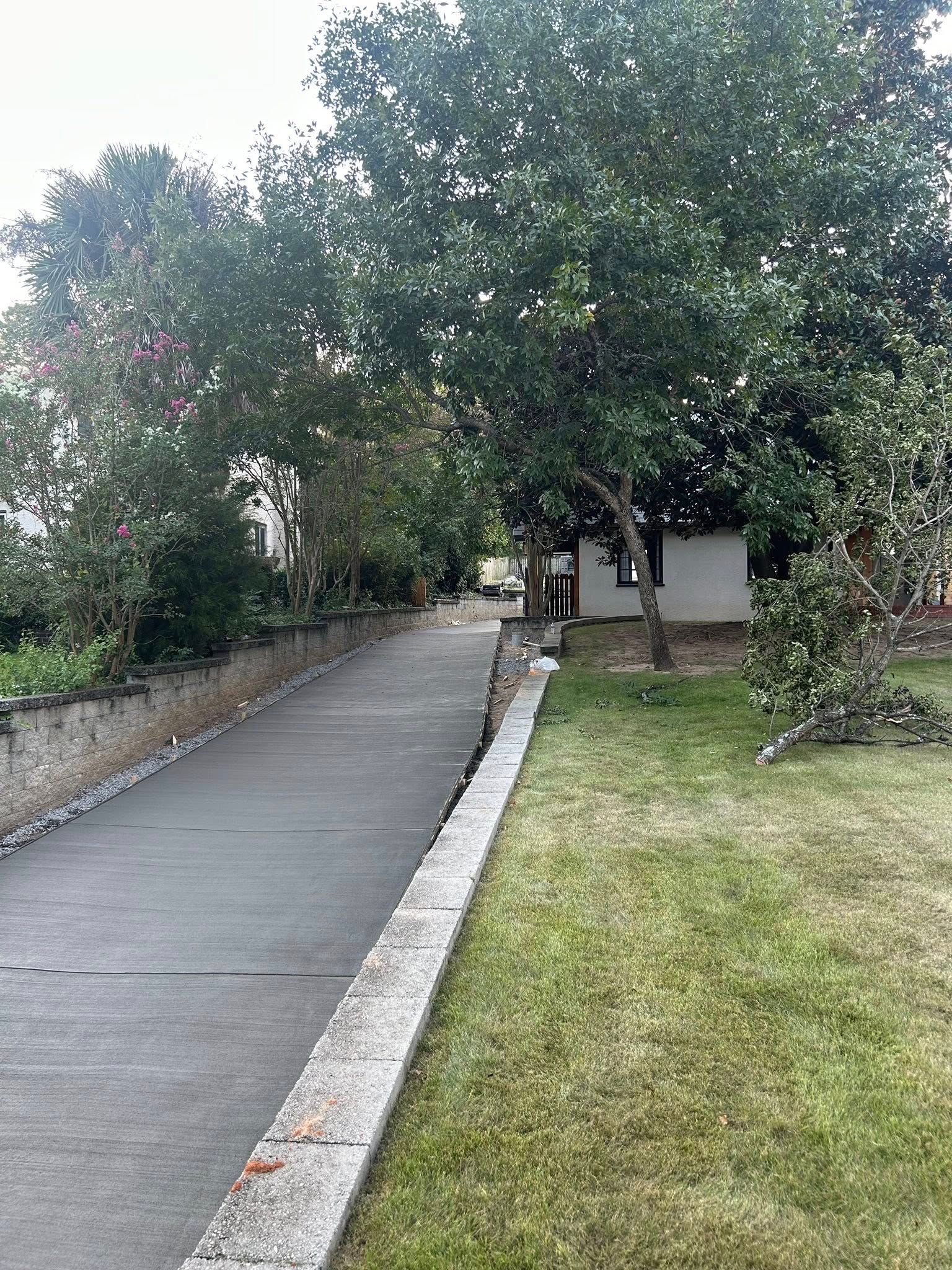 Concrete path alongside a stone wall and grass, leading towards a small building and trees.