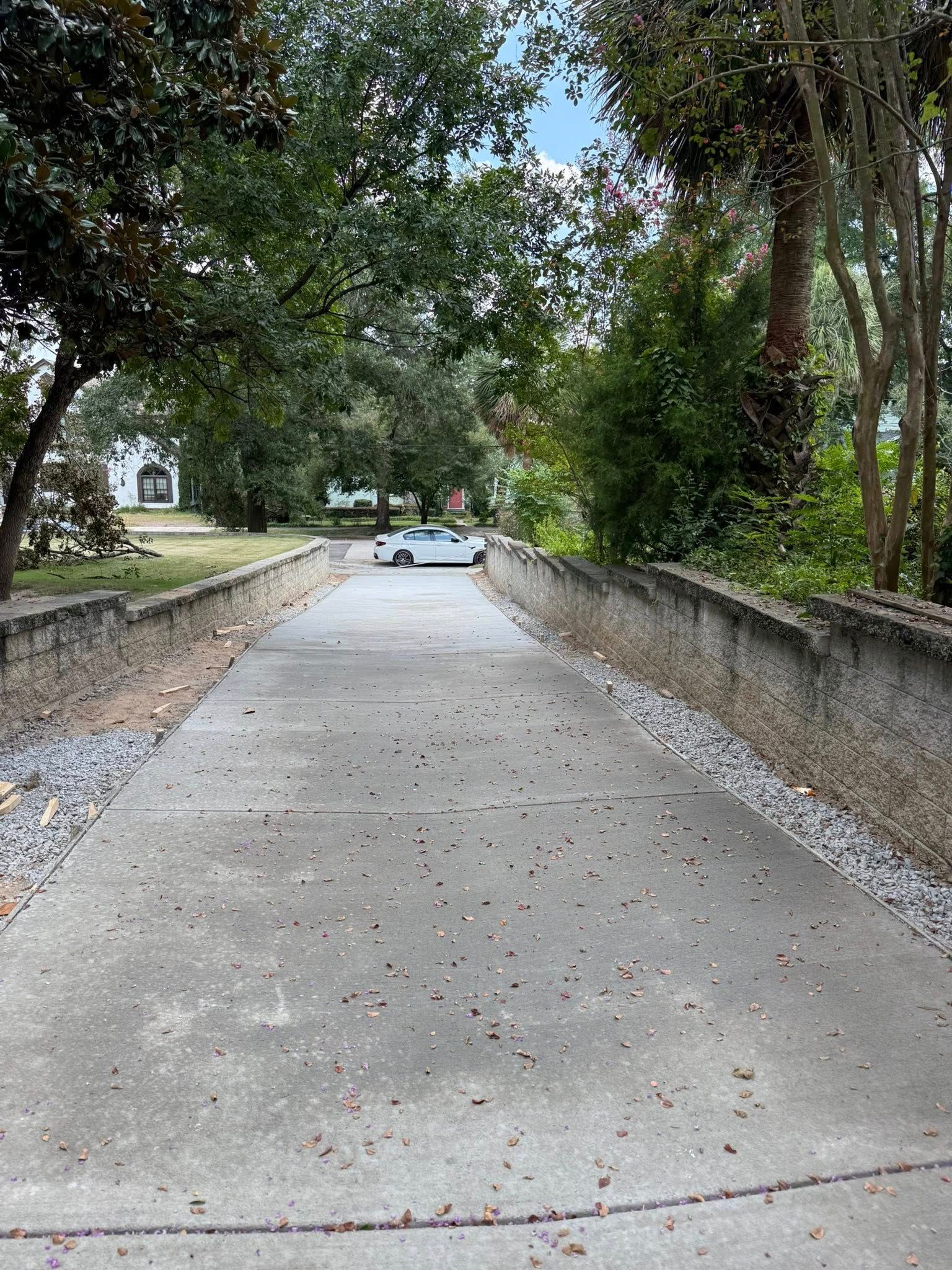 Concrete driveway lined with low walls, leading to a distant car and trees under a blue sky.