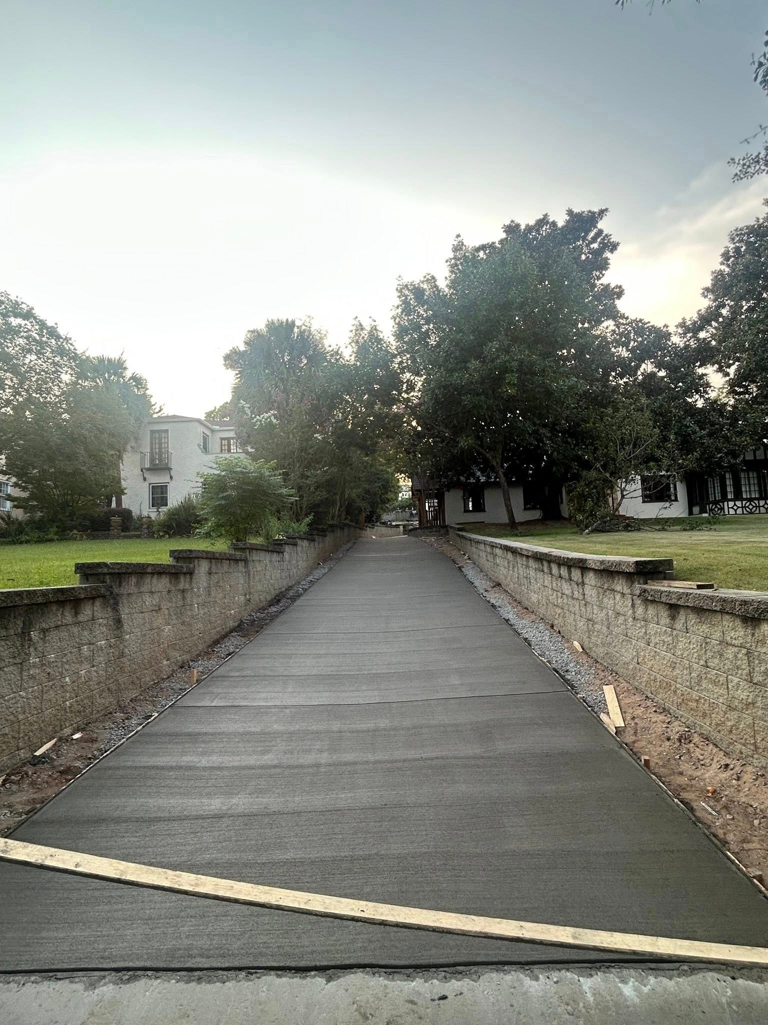 Asphalt path between stone walls, trees along sides, buildings in the distance, overcast sky.