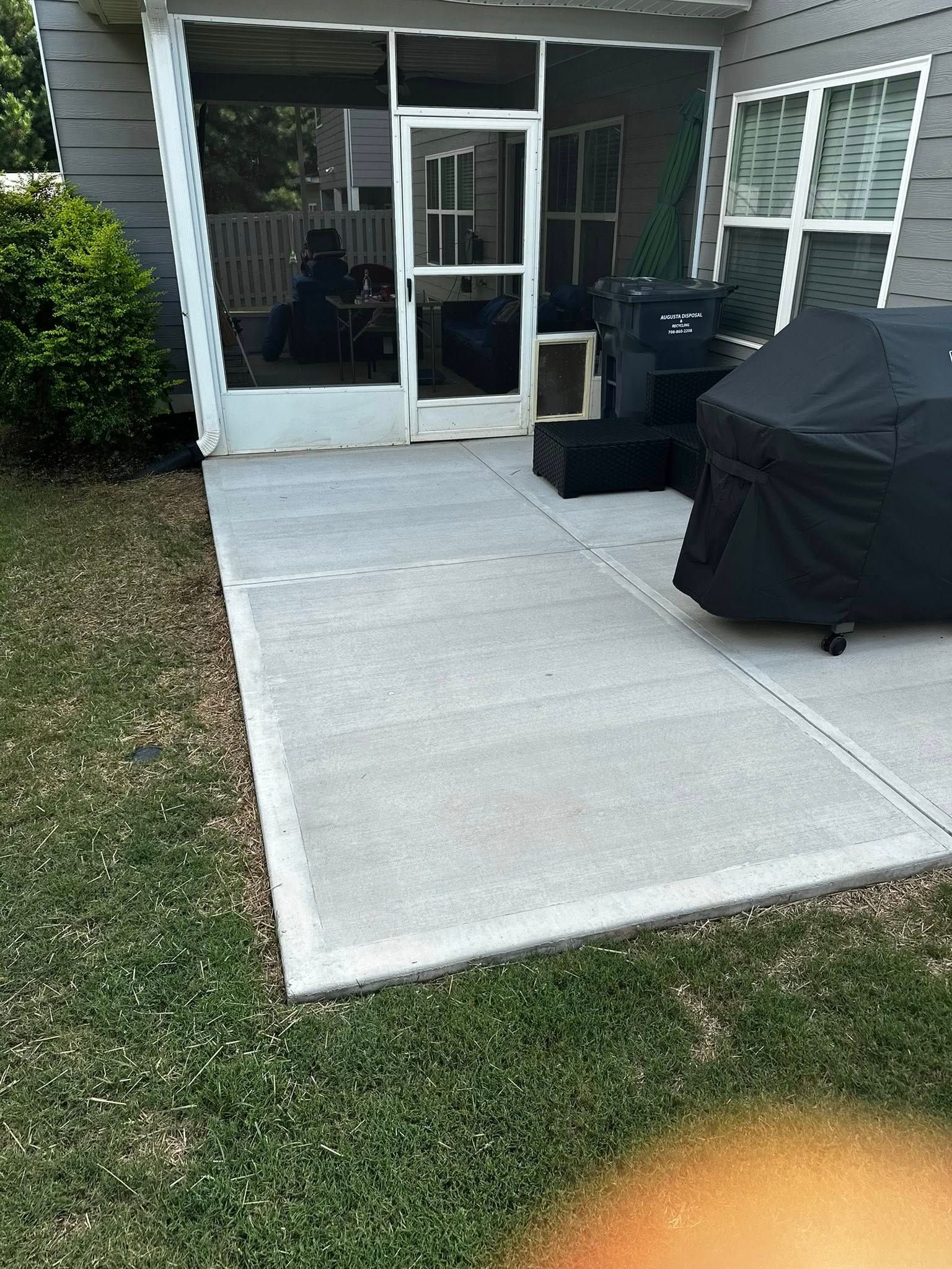 Concrete patio beside grass, leading to screened porch door. Grill covered in black; house in the background.