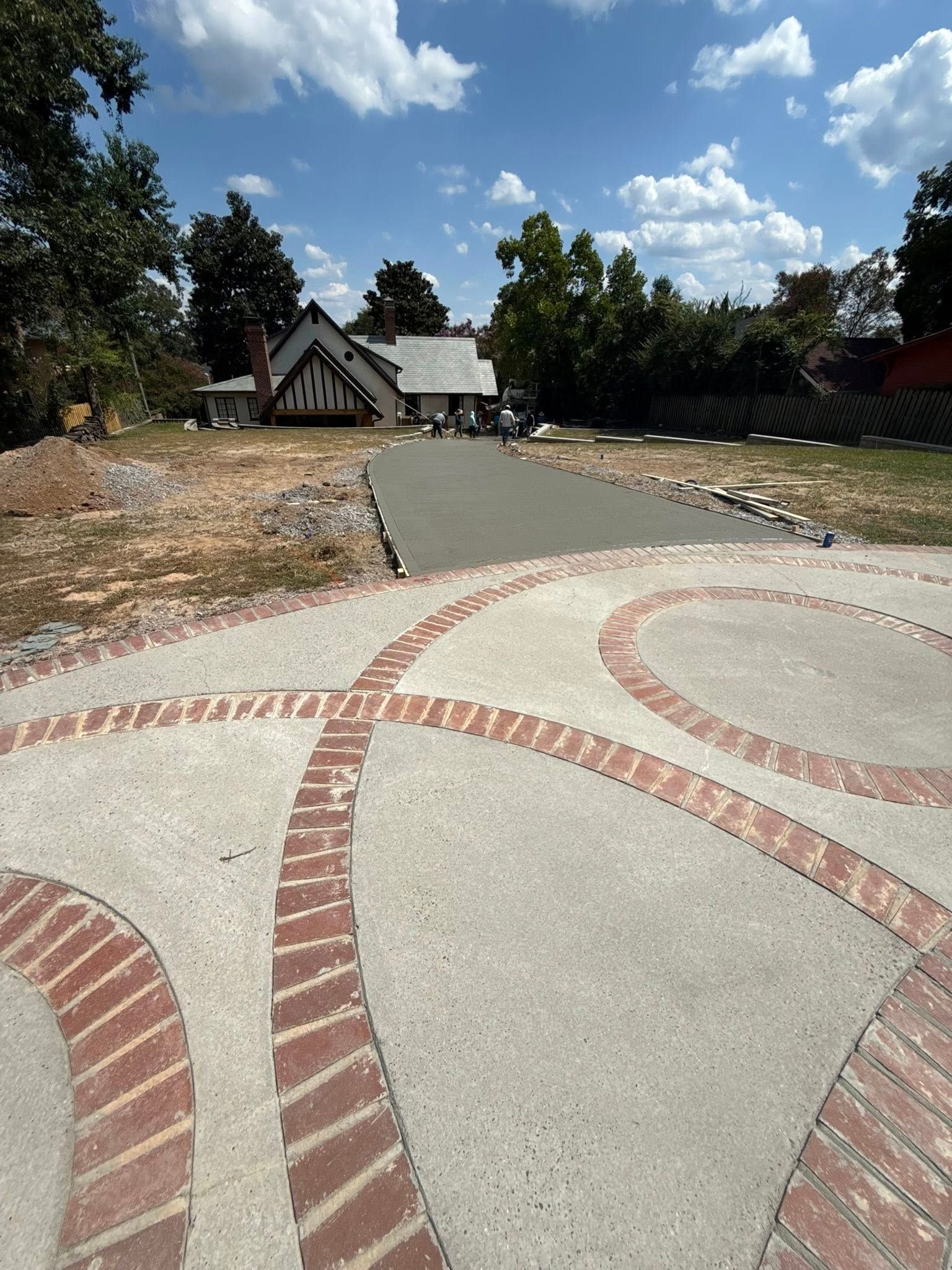 Concrete driveway with brick-lined patterns leading to a house, green trees and blue sky with clouds.