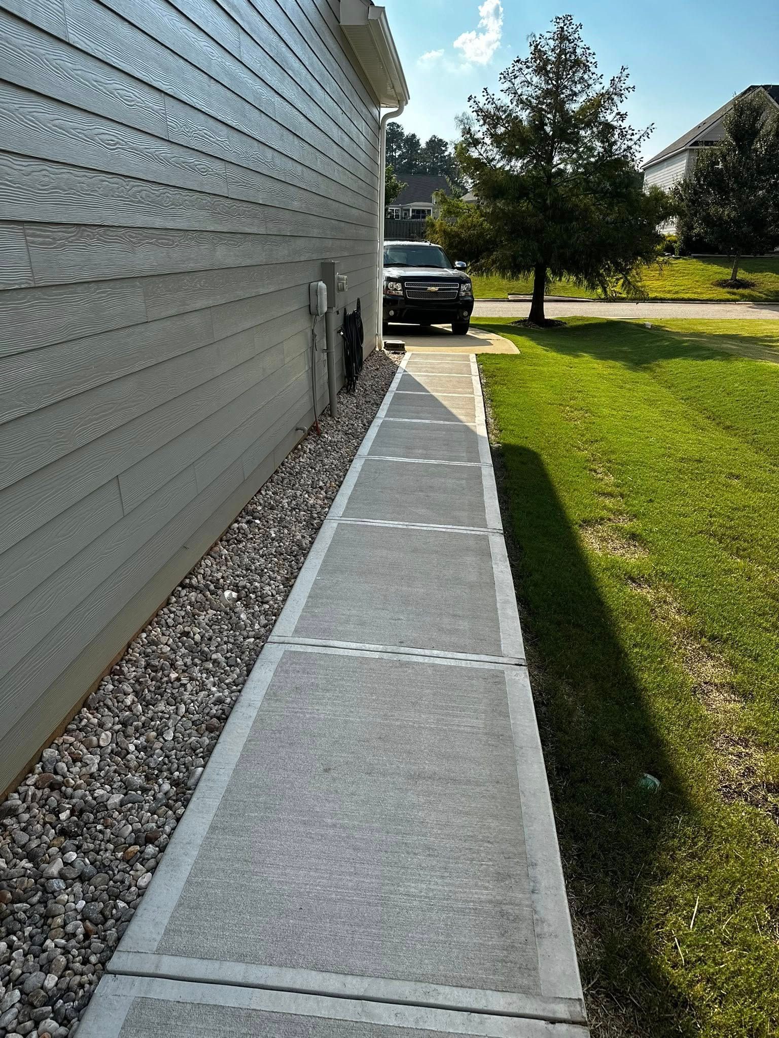Concrete sidewalk next to a light-colored house and gravel bed, leading to a parked truck. Sunny day.
