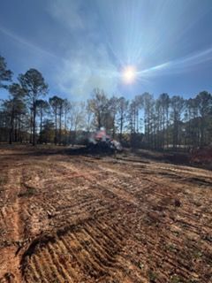 Clearing of a wooded area with burning debris and smoke under a sunny blue sky.