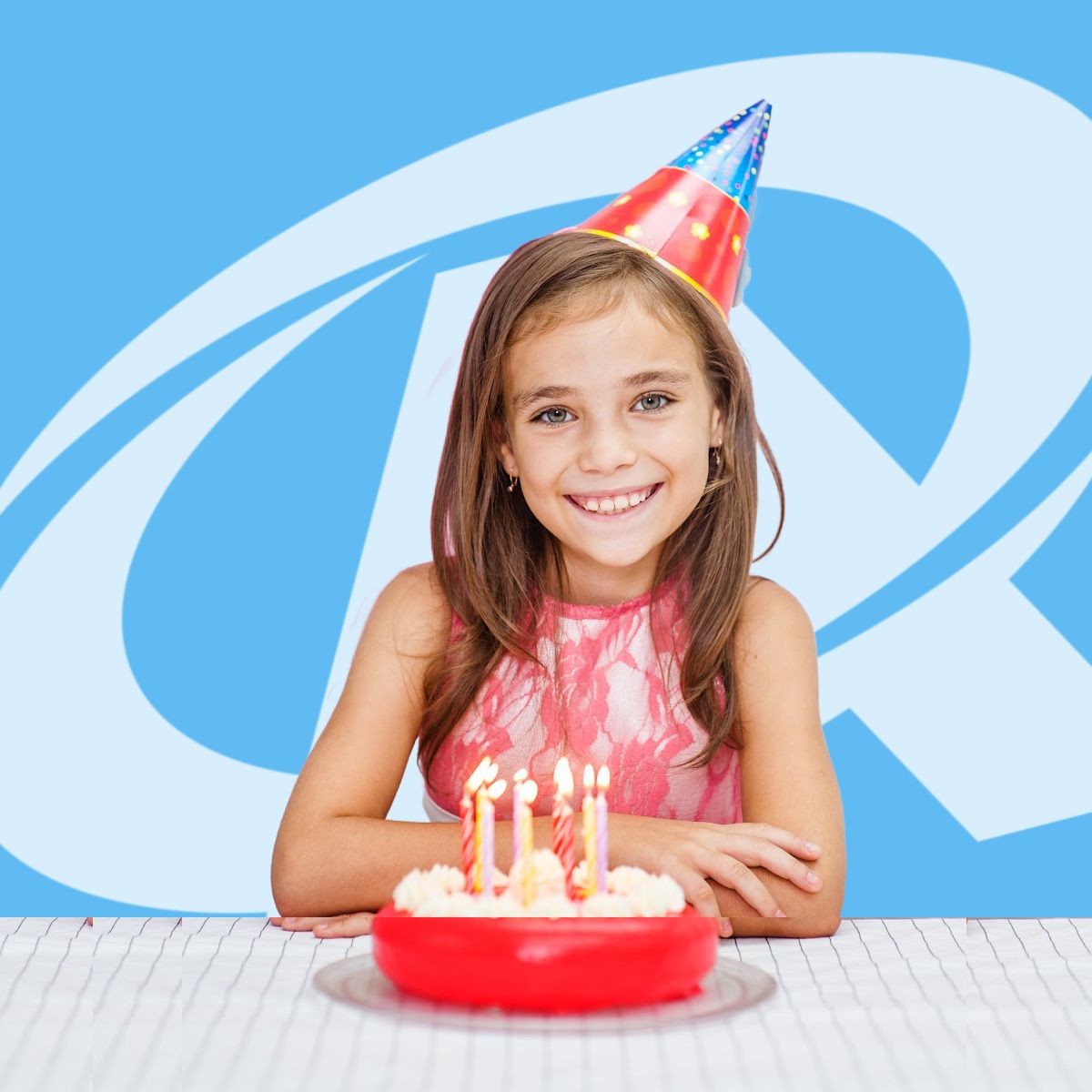 A little girl wearing a party hat sits in front of a birthday cake