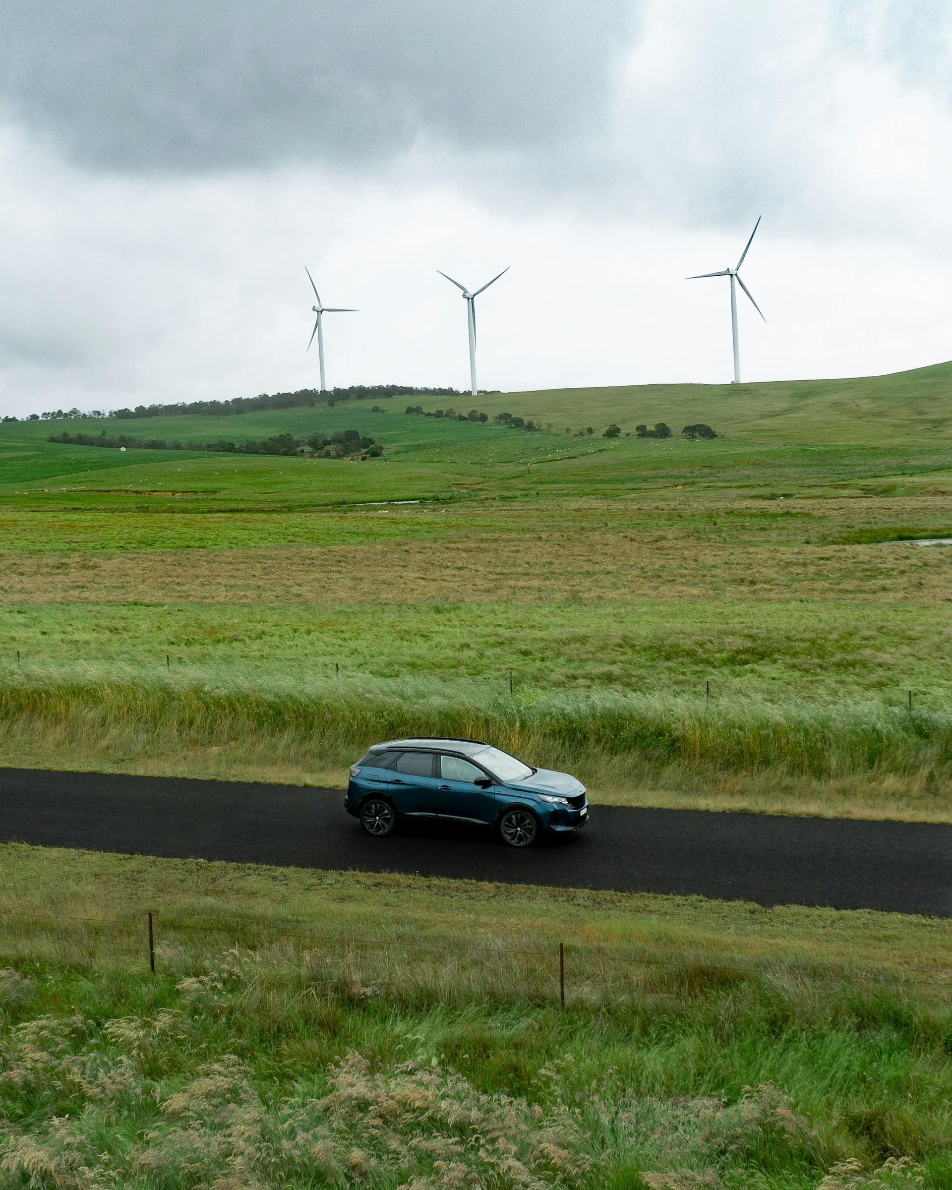 Blue car on a rural road with wind turbines on a grassy hillside under a cloudy sky