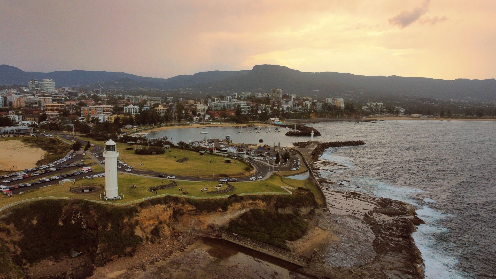 Coastal lighthouse and grassy park overlooking a bay at sunset, with city skyline and mountains in the distance