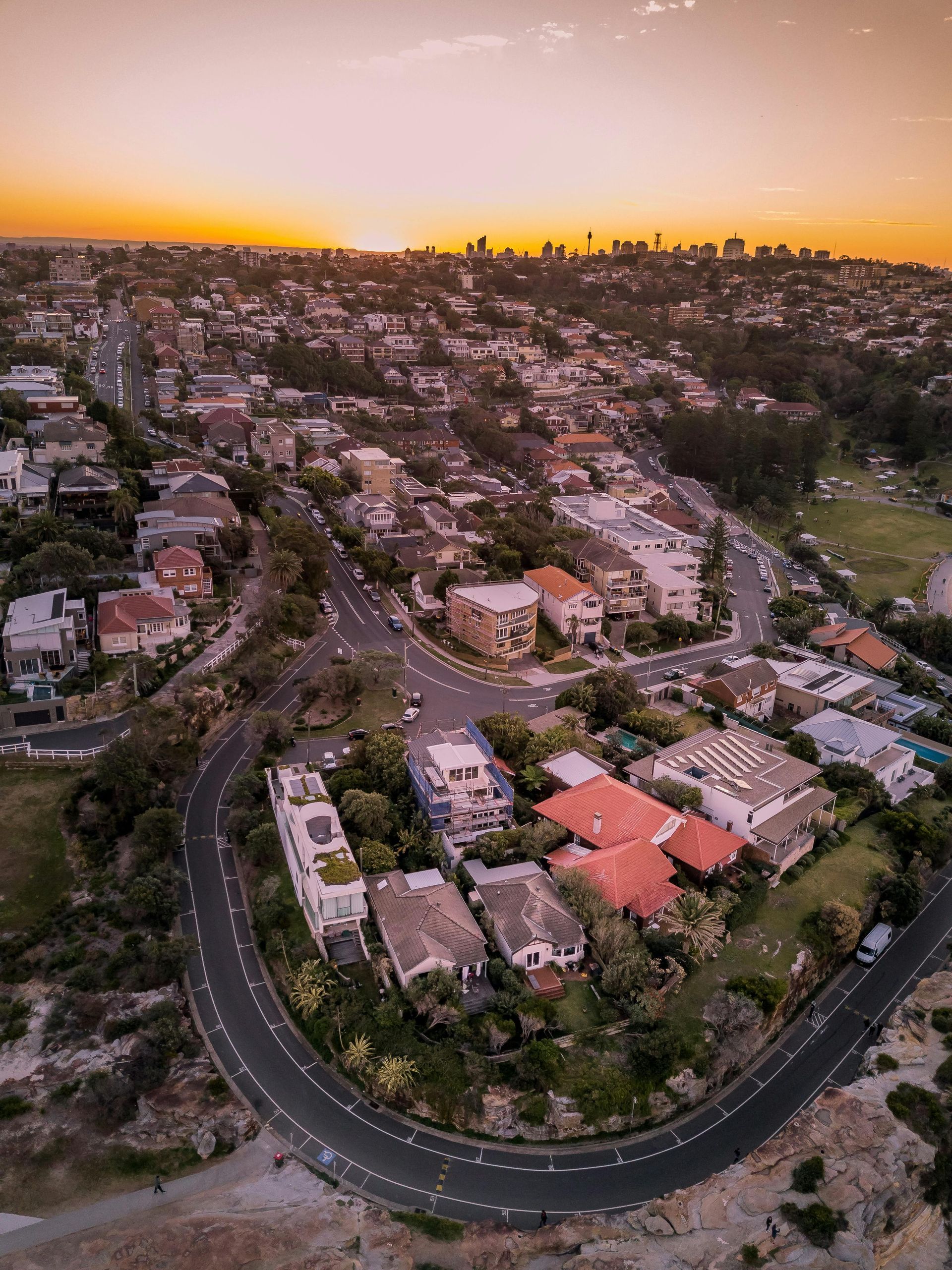 Aerial view of a suburban neighborhood at sunset with winding roads and a distant city skyline