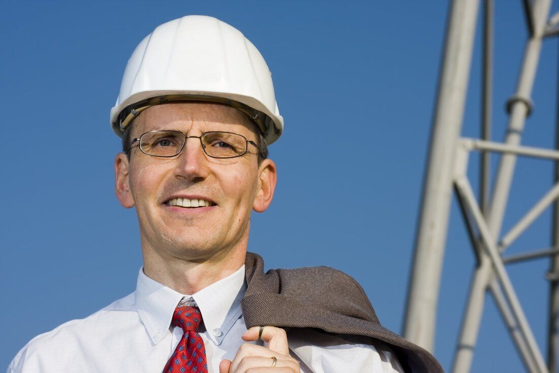 Smiling engineer in white hard hat and glasses, holding jacket over shoulder at a construction site.
