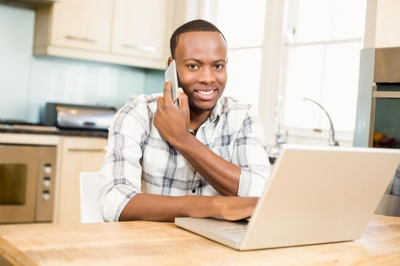 Man smiling at a laptop in a bright kitchen, talking on a phone at a wooden table