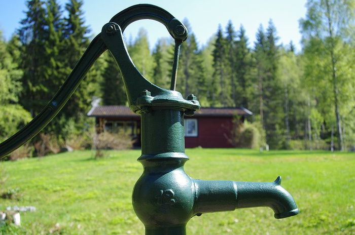 Green hand water pump in a grassy yard with trees and a house in the background