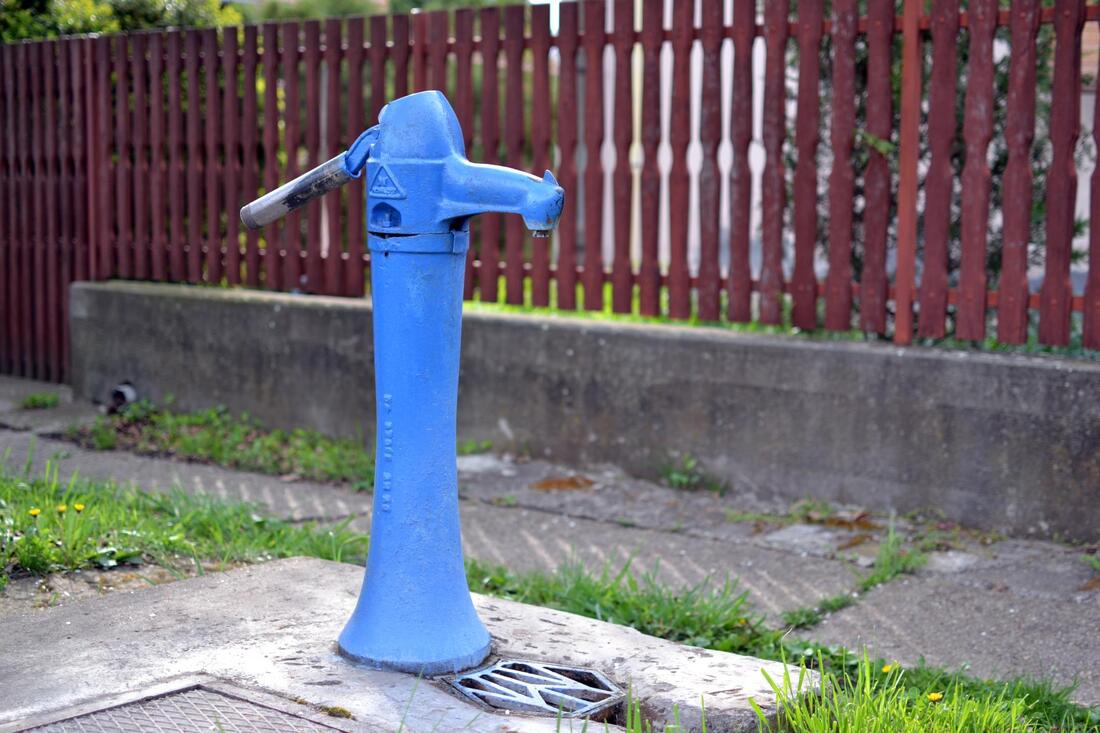 Blue water pump on a sidewalk beside a fence and concrete wall.