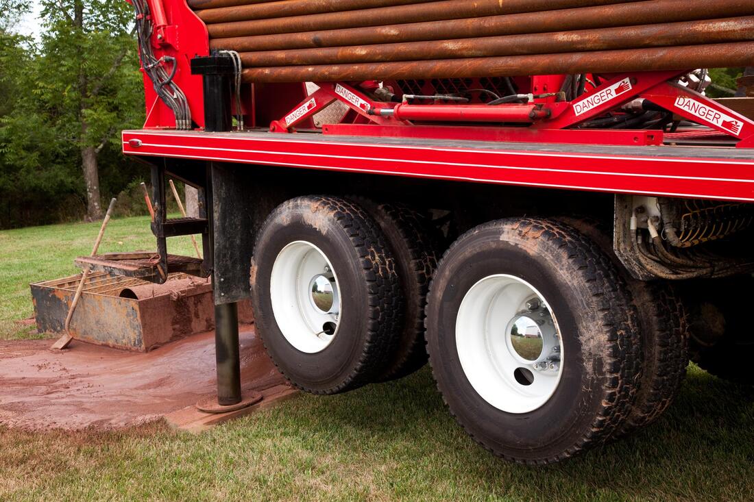 Red flatbed truck with dual rear wheels parked on grass, carrying wooden logs or pipes.