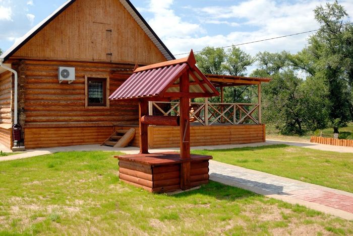 Wooden house with a red roof and a well in a grassy yard under a partly cloudy sky