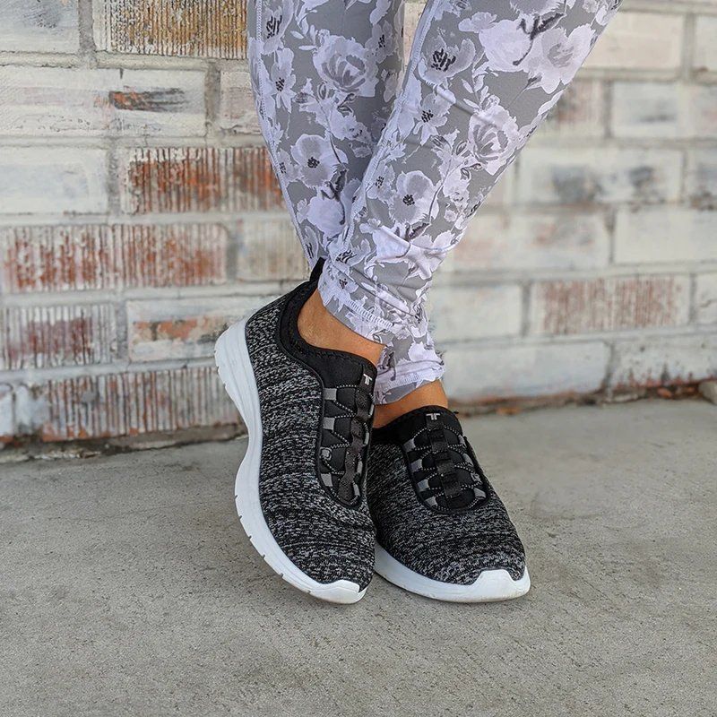 A woman wearing a pair of black and white sneakers and floral leggings is standing in front of a brick wall.