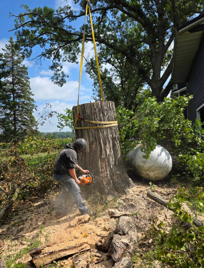 Man cutting tree trunk with chainsaw, lifted by a crane. Outdoors near a building and gas tank.