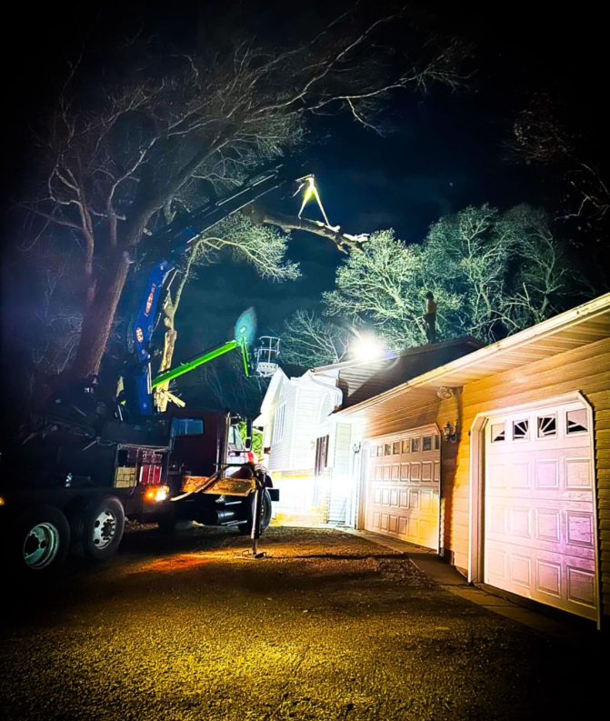A tree trimmer in a lift truck trims a tree over a house at night.