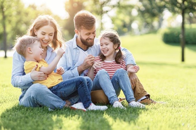 Family of four laughing together while sitting on green grass in a sunny park.