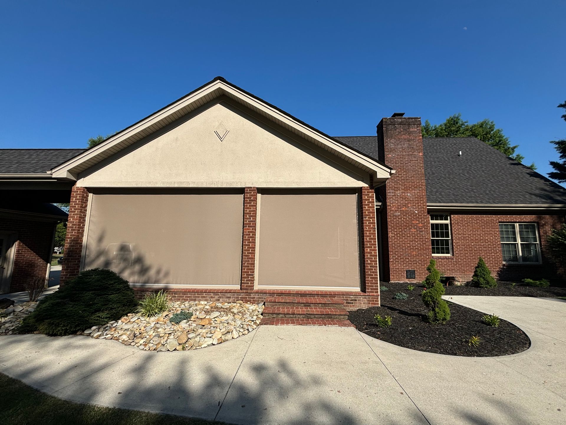 Tan retractable screens cover a porch, brick exterior, and a dark shingle roof against a blue sky.